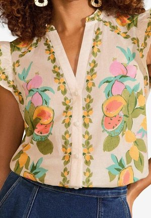 Vrouw draagt een mouwloze witte blouse met kleurrijke bloemen- en fruitprints, gecombineerd met hooggetailleerde blauwe jeans en ronde oorbellen.