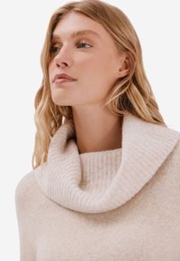 Beżowy sweter typu cowl-neck wykonany z miękkiego, teksturowanego materiału z prążkowanymi detalami przy kołnierzu. Gładkie wykończenie, luźny krój.