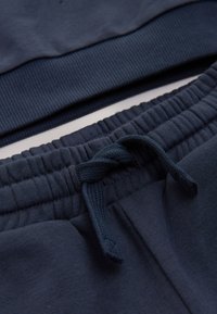 Gros plan sur la ceinture d'un pantalon de survêtement bleu foncé avec élastique et cordon noué, et l'ourlet assorti d'un sweat sur fond blanc.
