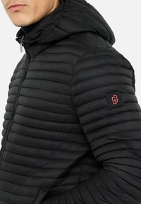 Svart puffjacka med quiltat mönster, huva, dragkedja och en logotyp-patch på ärmen. Har ett slätt, lätt material.