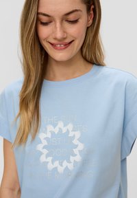 Lichtblauw katoenen T-shirt met korte mouwen, voorzien van een ontwerp in wit en goud in een artistiek cirkelpatroon.