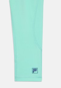 Τμήμα από αθλητικά κολάν σε ανοιχτό τιρκουάζ χρώμα με λείο υλικό, αντίθετη ραφή και μπλε λογότυπο στην κάτω δεξιά γωνία.