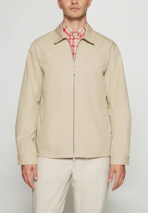 Mand iført beige lynlåstrøje over en rød og hvid ternet skjorte med matchende beige bukser mod enkel baggrund.