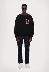 Zwarte oversized sweatshirt met een rode "F" patch, gecombineerd met donkerblauwe jeans en witte sneakers. Model draagt donkere zonnebril.