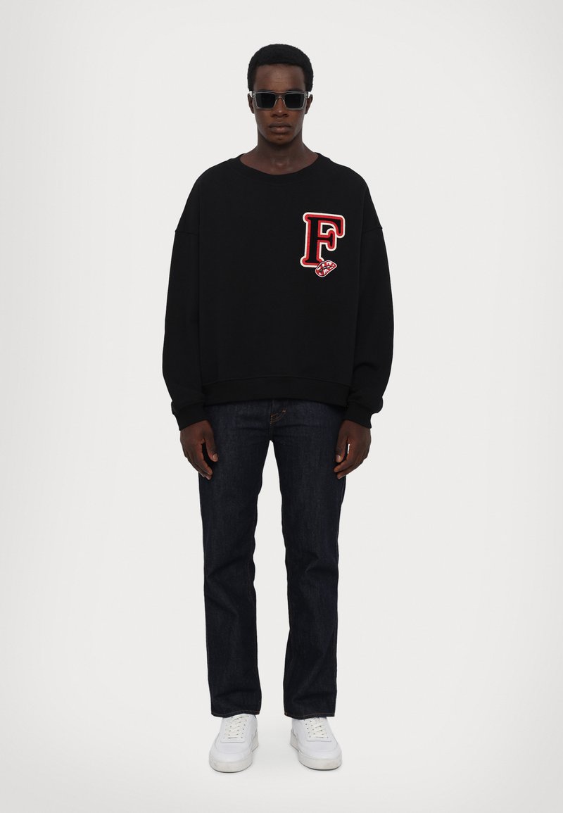 Zwarte oversized sweatshirt met een rode "F" patch, gecombineerd met donkerblauwe jeans en witte sneakers. Model draagt donkere zonnebril.