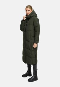 Žena oblečená v dlhom olivovo zelenom pufferi s kapucňou, čiernych legínach a masívnych čiernych čižmách, stojaca proti jednoduchému pozadiu.