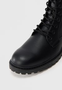 Stivaletto in pelle nera con superficie testurizzata, punta tonda e design con lacci. Presenta una suola in gomma resistente con scanalature per una migliore aderenza.