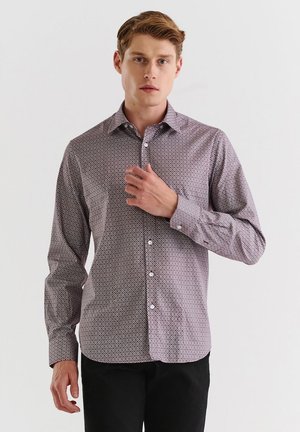 Koszula z długim rękawem w purpurowo-białym geometrycznym wzorze; zapięcie na guziki z przodu; kołnierz; miękka, gładka tekstura. Model pozował z ręką na kołnierzu.