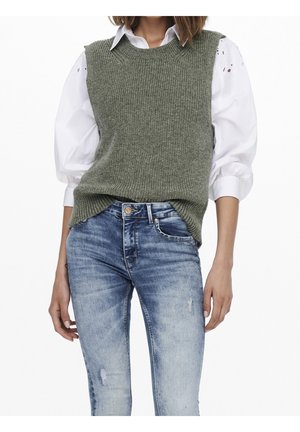 Femme portant un pull sans manches vert en tricot sur un chemisier blanc à manches bouffantes, associé à un jean bleu délavé, debout devant un fond blanc.
