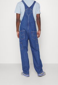 Džíny s laclem ve středně modré barvě s nastavitelnými popruhy, dvěma předními kapsami a dvěma zadními kapsami. Volný střih, pevná látka a rovné nohavice.