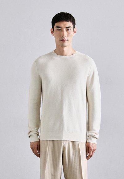Pull en maille beige clair avec un col rond et des manches longues, présentant un motif texturé et une coupe décontractée. Associez-le à un pantalon léger à rayures.