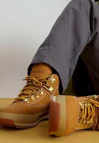 Botas de senderismo Timberland en cuero color marrón, con cordones amarillos, ojales de metal y una suela de goma marrón resistente. Pantalones grises son parcialmente visibles.