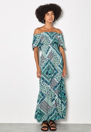 Donna con acconciatura afro che indossa un vestito lungo a spalle scoperte con motivi nei colori teal, blu e bianco, in piedi contro un muro semplice.