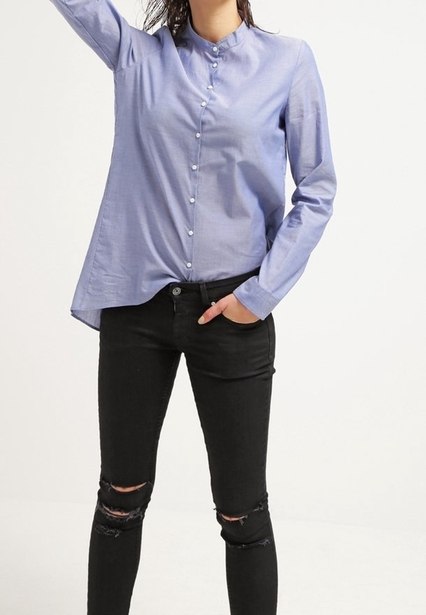 Personne portant une chemise bleu clair boutonnée avec un col montant et un jean skinny noir déchiré, une main dans la poche et l’autre levée.