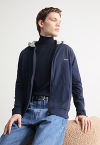 Felpa con zip di colore blu navy con accenti grigi, realizzata in misto cotone, con tasche laterali, indossata sopra un dolcevita nero, abbinata a jeans blu.