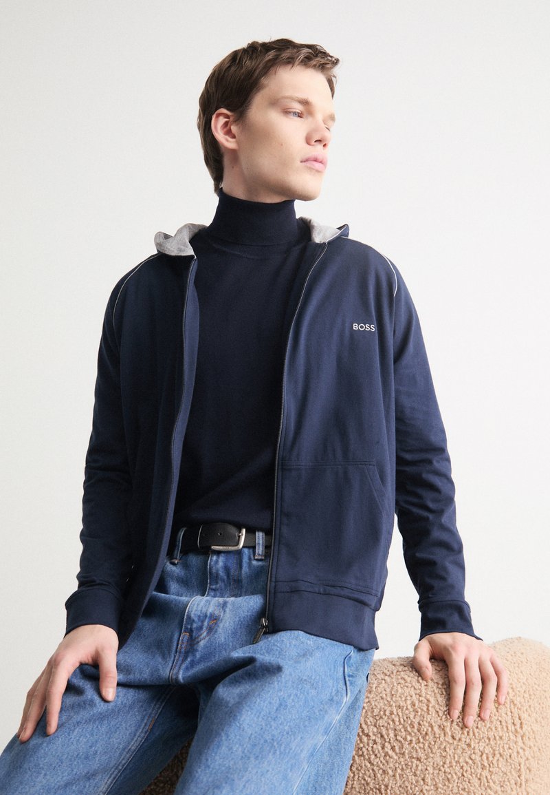 Felpa con zip di colore blu navy con accenti grigi, realizzata in misto cotone, con tasche laterali, indossata sopra un dolcevita nero, abbinata a jeans blu.