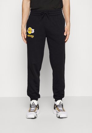 Uomo con pantaloni della tuta neri Los Angeles Lakers e scarpe sportive bianche con dettagli neri e dorati, in piedi davanti a un muro semplice.