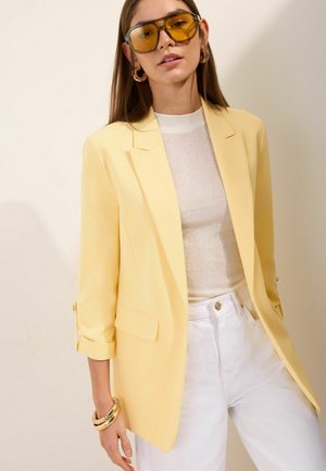 Woman wearing oversized yellow sunglasses, yellow blazer, white top, white pants, and gold hoop earrings and bracelets against plain background.