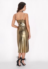Rochie din satin auriu metalizat cu bretele subțiri, formă ajustată și lungime până la genunchi. Asortată cu pantofi cu toc negri, cu accente de pene.