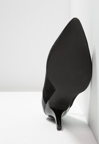 Chaussure à talon haut noire à bout pointu avec dessus en suède et détail en cuir texturé, photographiée de la semelle vers le haut sur fond blanc.