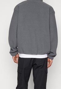 Giacca in pile grigia con superficie texturizzata, colletto alto e vestibilità oversize. Presenta polsini e orlo a coste, indossata sopra una camicia bianca.