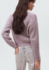 Pleteni pulover v mehki mauve barvi, z teksturiranim vzorcem, spuščeni rameni in rebrastim robom, v kombinaciji s svetlosivimi kavbojkami.