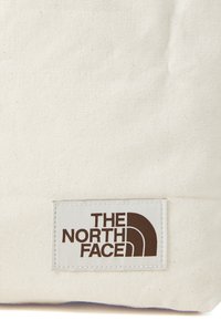 Šviesiai grietininis medvilninės priekinės rankinės maišelis su tekstūruota apdaila. Ant stačiakampio ženklo matomas "THE NORTH FACE" logotipas ruda spalva.