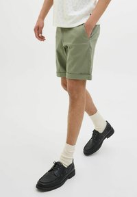 Olive green shorts with rolled hem, paired with black leather shoes and cream socks. The fabric appears smooth and sturdy.