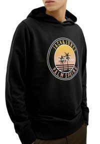 Sudadera negra con un gráfico frontal que presenta palmeras y colores de atardecer. El logo redondeado incluye el texto "JACK & JONES" y "PALM SPRING".