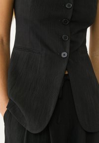 Černá přizpůsobená vesta s texturovanou látkou, vybavená šesti knoflíky, slim fit a decentním výstřihem do V. Obsahuje boční kapsy.