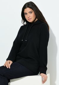 Sudadera negra con capucha hecha de tela suave, que presenta un ajuste holgado, puños acanalados y cordones con acentos metálicos. Combinada con unos pantalones vaqueros azul oscuro.