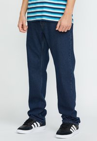 Jeans in denim blu scuro con design a gamba dritta, caratterizzati da cuciture a contrasto, stile cinque tasche e indossati con sneakers nere con strisce bianche.