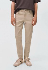 Jeans de algodón beige con corte ajustado, que cuentan con cinco bolsillos y cierre de botón. Se combinan con zapatillas ligeras y una camisa oscura.