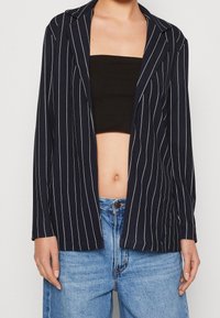 Námořnické pruhované sako s tenkými bílými proužky, dlouhými rukávy, klopovým límcem a otevřeným předním dílem. Kombinováno s černým crop topem a džíny s vysokým pasem.