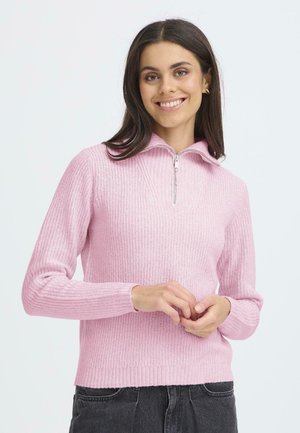 Lys pink strikket sweater med kvart-zip krave, ribbet tekstur, lange ærmer og en tætsiddende kant, båret over mørke jeans.