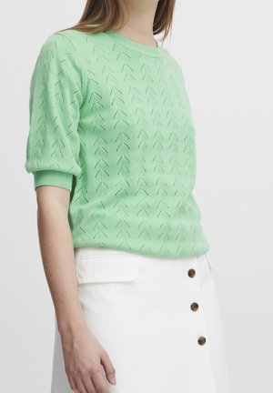 Světle zelený pletený svetr se strukturovaným vzorem šipek, kulatým výstřihem a krátkými rukávy. Kombinován s bílou sukní s knoflíkovými detaily.