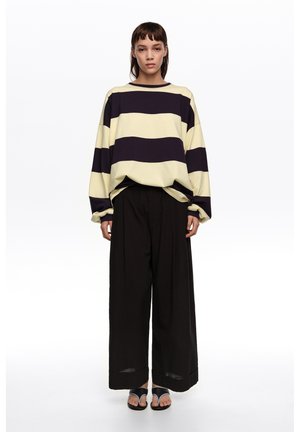 Mujer con una blusa de manga larga suelta a rayas negras y beige, pantalones anchos negros y sandalias negras, de pie contra un fondo blanco.