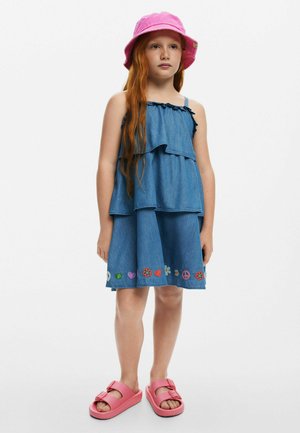 Robe en denim avec des volants superposés, ornée d'accents floraux brodés de différentes couleurs le long de l'ourlet. Portée avec un chapeau seau rose et des sandales douces.