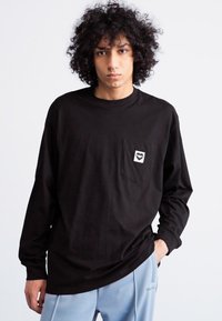 Black long-sleeve cotton t-shirt with a front pocket and a small heart graphic. Model wears light blue pants, showcasing a relaxed fit.