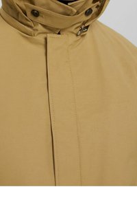 Beżowy płaszcz z wysokim kołnierzem, zapięciem na guziki i gładką teksturą. Widoczne przeszycia na przodzie i kołnierzu.