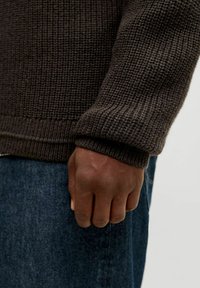Dunkelbrauner gestrickter Pullover mit einem strukturierten Muster, ausgestattet mit einem gerippten Bündchen und Saum, kombiniert mit dunklen Jeans.