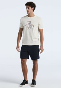 Lichte crèmekleurige t-shirt met een pinguïnafbeelding in rood, blauw en zwart; gecombineerd met marineblauwe shorts. Model draagt zwarte sneakers.