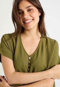 Top verde oliva con escote en V y mangas cortas, que presenta una tela texturizada y cinco botones de madera en la parte delantera. Diseño simple y casual.