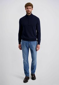 Navy Zip-Pullover aus gestricktem Material, kombiniert mit hellblauen Denim-Jeans und dunkelbraunen Schuhen, vor einem einfarbigen Hintergrund stehend.