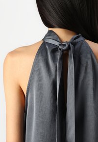 Haut halter gris avec une finition texturée, présentant une fermeture au cou nouée à l'arrière et un design à dos ouvert. Le tissu lisse tombe élégamment.