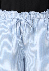 Spodnie w pionowe paski w jasnoniebieskim i białym kolorze, z falbaną na elastycznym pasie i zawiązywanym sznurkiem, noszone z czarną bluzką.