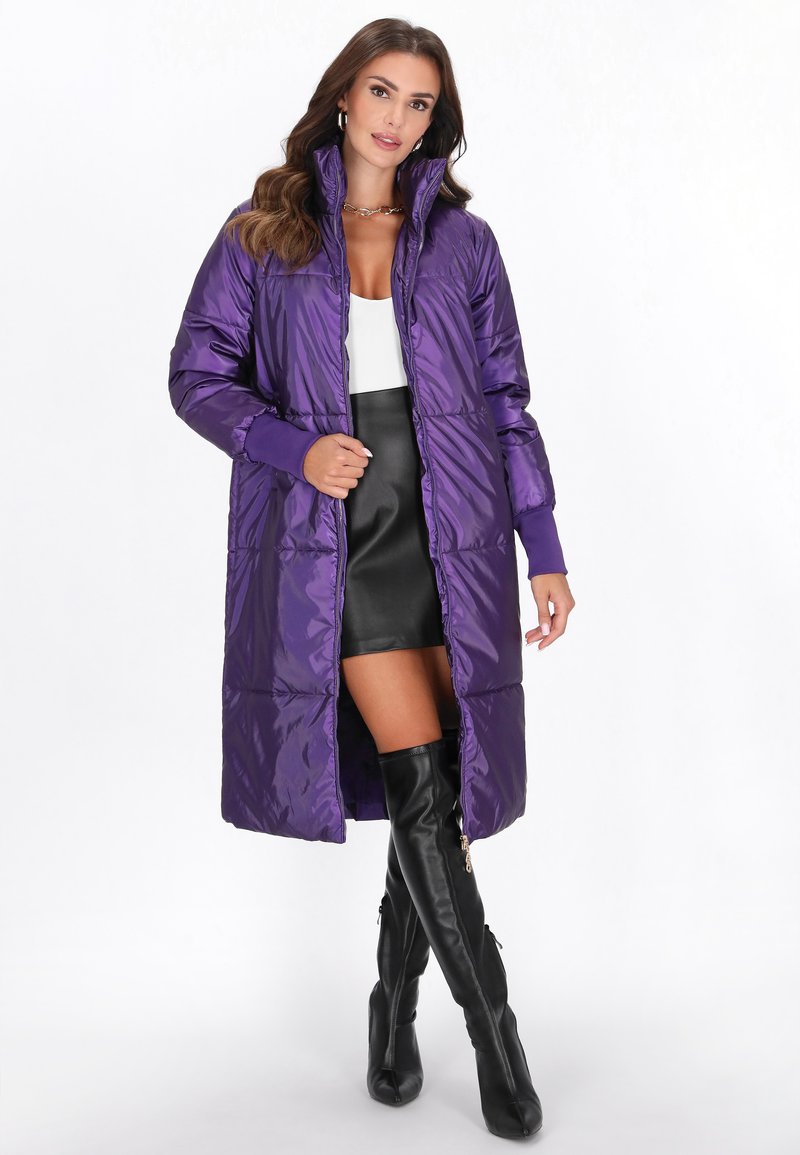 Cappotto lungo piumino viola con colletto alto, chiusura a zip frontale e polsini a costine. Presenta una finitura lucida e un design trapuntato. Indossato sopra una minigonna nera.