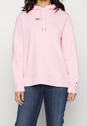 Mujer vistiendo una sudadera con capucha de color rosa claro con el texto "Hilfiger" y cordones, combinada con vaqueros azul oscuro.
