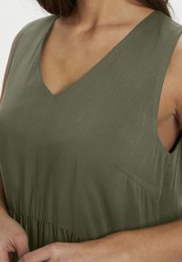 Robe sans manches de couleur vert olive avec un col en V, une taille froncée et une texture subtile. Comprend une petite poche sur le côté.