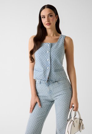 Femme aux longs cheveux foncés portant un haut sans manches texturé bleu clair et un pantalon assorti, tenant un sac à main structuré blanc.